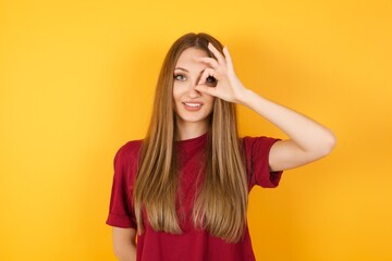 Fototapeta premium Beautiful Young beautiful caucasian girl wearing red t-shirt over isolated yellow background doing ok gesture with hand smiling, eye looking through fingers with happy face.