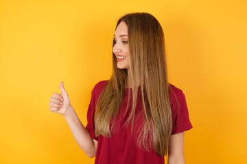 Beautiful Young beautiful caucasian girl wearing red t-shirt over isolated yellow background Looking proud, smiling doing thumbs up gesture to the side. Good job!
