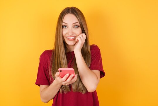 Beautiful Young beautiful caucasian girl wearing red t-shirt over isolated yellow wall poses with mobile phone device, types text message on modern smartphone, watches funny video during free time, 