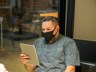 Shot of a man in a medical mask in a cafe  looking at his  tablet - new normal