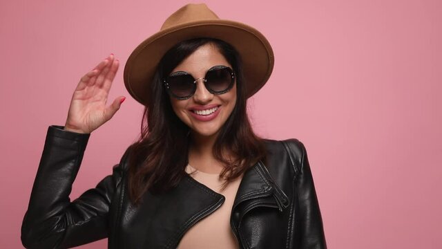 smiling cool woman in leather jacket wearing sunglasses and hat, laughing, fixing hat, looking down and touching forehead in a shy manner, smiling, crossing arms and posing on pink background