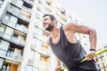 Young man exercising outside. Cheerful positive sportsman holding hands on sporty bar and keep his upper body above it. Training strenghts of his body. Exercising at urban buildings.