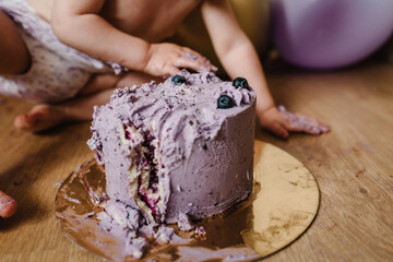 Little cheerful baby girl with the first cake to birthday on balloons background. Smash cake. Funny toddler eating cake. Dirty sticky hands from crumb pie.
