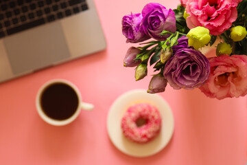 Girly workspace with flowers, laptop, donut, and coffee on pink background. Morning and breakfast concept. Selective focus, blurred background. Girly stuff, cute working space. Top view, flat lay