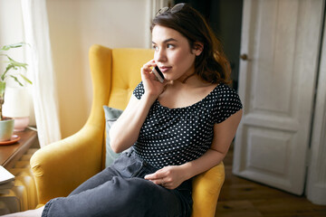 Attractive young European woman sitting comfortably in yellow armchair making phone call. Pretty...