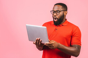 Young surprised shocked amazed african man standing and using laptop computer isolated over pink...