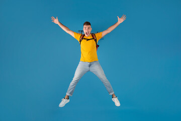 Fototapeta na wymiar Handsome millennial guy with backpack jumping in excitement over blue studio background