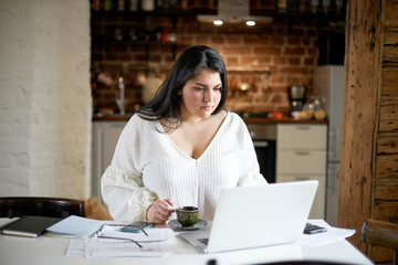 Serious plus size young businesswoman in white blouse working distantly from home office checking...