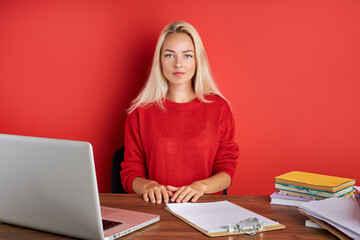 portrait of young caucasian blonde female at work place, she sits in casual wear at office desk, enjoy working on laptop and with documents. isolated red background