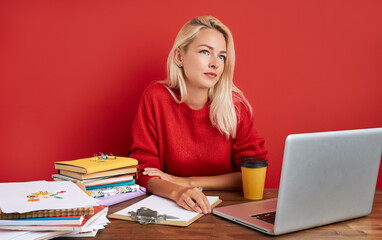 portrait of confident caucasian blonde female sitting at office desk with laptop and papers, young lady in casual wear work as manager, has too much work, think, isolated over red background