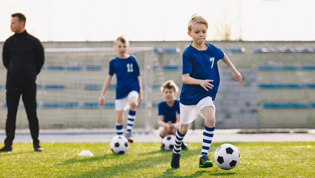 Group of School Kids with Young Coach Kicking Soccer Balls on Grass Sports Pitch. Junior Footballers in Blue Shirts on Training. Horizontal Image of Soccer Club Practice