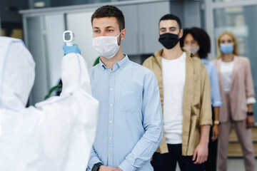 Obraz na płótnie Canvas Man in protective suit measures temperature of workers in protective masks in office interior, stopping spread of covid-19
