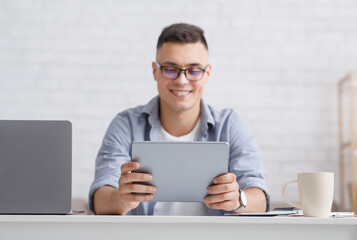 Man looking at web camera making conference video call. Smiling teacher holds digital tablet, sitting at table with laptop in living room interior