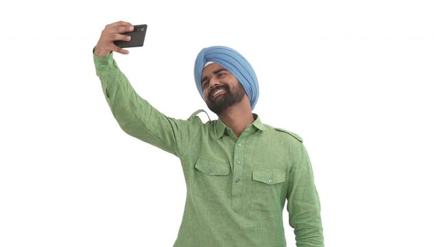 Young Indian man taking selfie with smartphone. Guy posing for camera, sending air kiss, showing V-sign and sticks out tongue. Isolated on white background