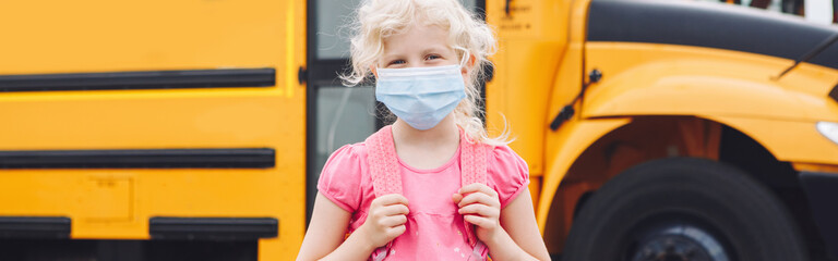 Girl student in face mask near yellow bus. Kid with personal protective equipment on face....
