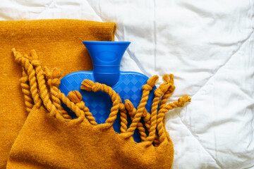 Eine blaue Wärmflasche in eine gelbe Wolldecke gewickelt auf einer weißen Bettdecke. Winter,...