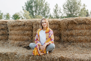 Naklejka na ściany i meble Autumn fall woman portrait. Portrait of beautiful Caucasian middle age woman sitting on haystack on farm. Pensive woman wrapped in yellow scarf blanket in park outdoors. Fall colors.