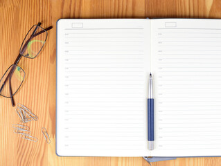 The diary open on a blank page lies on a wooden table. Nearby there are a pile of paper clips and glasses. There is a ballpoint pen on the diary.