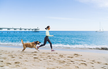 Side view of satisfied female athlete running at seashore with mongrel dog feeling happiness and...