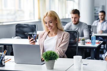 Manager works with client online. Smiling woman worker holding smartphone and typing on laptop at workplace in modern office interior