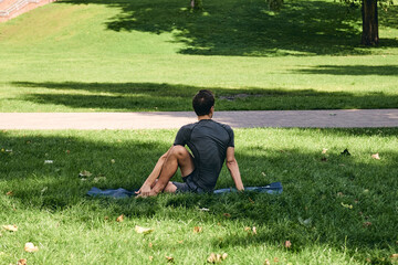 Naklejka na ściany i meble Young athletic man in sportswear doing yoga in the park. Practice half spinal twist asana outdoors. People exercising stretching on green grass with yoga mat. Fitness and healthy lifestyle