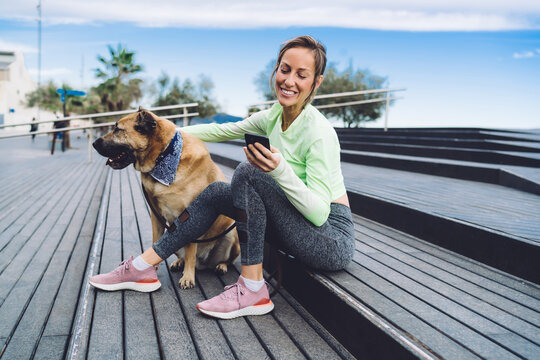 Prosperous female athlete in stylish tracksuit sitting at urbanity with mongler dog and laughing during cellphone communication, carefree fit girl enjoying friendly mobile messaging and networking