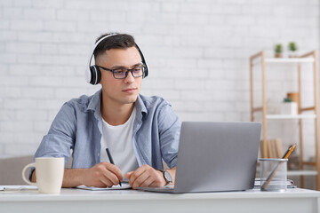 Modern student or pupil studies at home. Serious guy with glasses and headphones makes notes in...