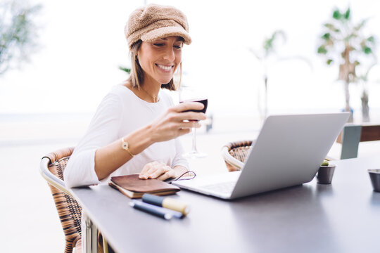 Smiling beautiful caucasian female watching video on laptop computer sitting on cafe terrace with glass of wine, positive woman blogger making video call in vlog enjoying social networks communication - Powered by Adobe