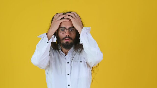 Shocked bearded young guy with long hair and white shirt surprised puts his hands to his head. Stressed and depressed guy isolated on yellow studio background