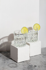 Vodka with ice and lime in two glasses on white stand. Shadows and sunlight trendy