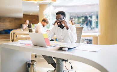 Obraz premium Happy dark skinned male manager satisfied with working process making smartphone call supporting clients, prosperous man employee satisfied with mobile phone talk using laptop computer in office