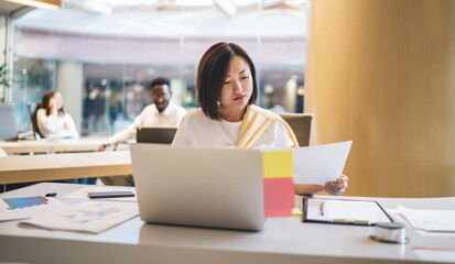 Concentrated asian female marketing manager holding blank paper documentation planning report for business, serious woman employee reading letters and mails checking plan for startup project in office