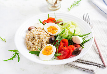 Breakfast oatmeal porridge with greek salad of tomatoes, cucumbers, olives and eggs. Healthy balanced food.