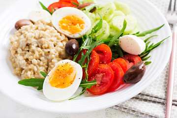 Breakfast oatmeal porridge with greek salad of tomatoes, cucumbers, olives and eggs. Healthy balanced food.