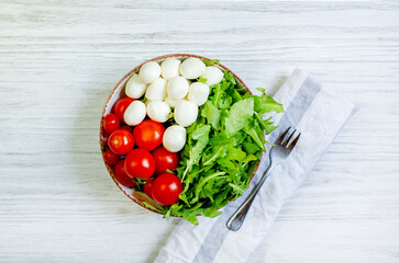 vegetable salad of tomatoes cherry peppers greens arugula mozzarella cheese in a round plate next to a fork on a white wooden table top view