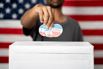 Close up of Hands dropping multiple I Voted sticker inside Ballot box with US flag as background,...