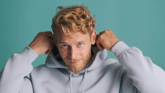 Handsome stylish blond bearded guy happily putting on and out a hood over colorful background