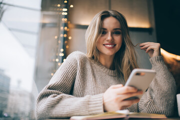 Portrait of cheerful hipster girl with perfect veneers smiling at camera while using smartphone...