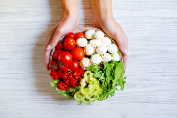 plate with vegetables tomatoes pepper arugula cheese mazzarella in female hands on a white wooden table top view