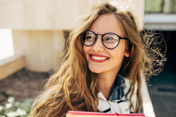 Closeup portrait of a smiling young student woman wearing transparent eyeglasses standing next to the college campus and carrying lots of books and folders on a sunny day. - obrazy, fototapety, plakaty
