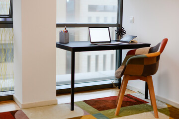 Office with a work table with a laptop, telephone, and pens, near a window, with a chair and a colorful carpet.