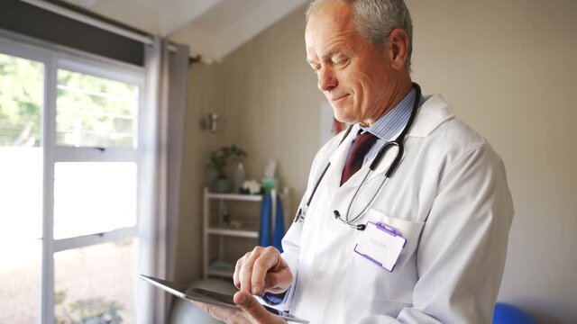 Elderly male doctor scrolling on digital tablet looking at outcomes of patient standing in room.