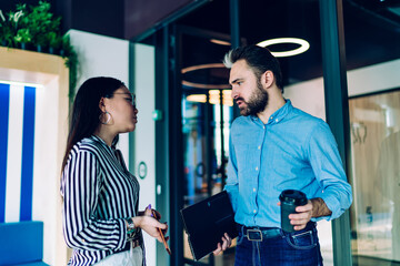 Diverse adult entrepreneurs looking at each other and talking while standing against glass door in office