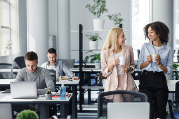 Gossip and coffee break at work. European and african american women with cups smile and talk in office interior with colleagues at workplaces with laptops and antiseptics