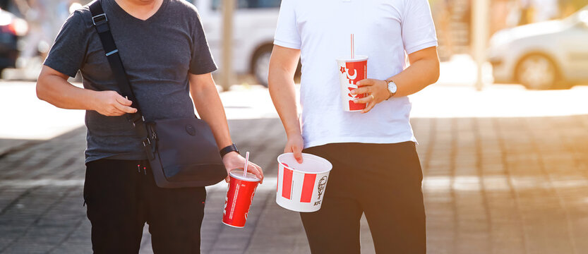 Minsk, Belarus. Sep 2019. Couple of men with Coca-Cola soda drinks eating KFC fried chicken from bucket while walking on street. Men has breakfast with fast food. Takeaway food. Hunger concept
