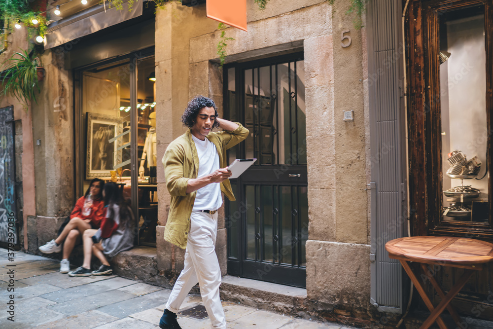 Poster handsome curly hipster guy in casual wear walking on urban settings using application on digital tab