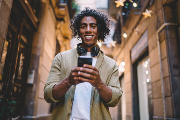 Portrait of cheerful handsome male with headphones walking on city street using mobile phone for browsing and reading information from social networks, smiling man millennial holding smartphone