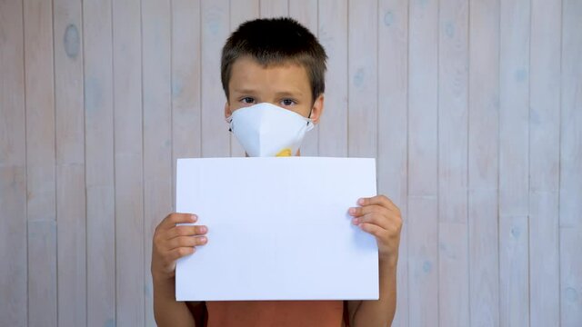 Little latin boy in a medical mask looking at the camera holding a sheet of a4 paper. Mockup in the hands of a child. Stay at home concept. Respiratory protection. hildren and Covid-19, Coronavirus