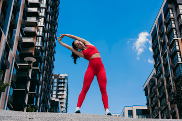 Beautiful woman with dark wavy hair wearing red sports top and leggins, white sneakers stretching arms. Sport in big city concept. Apartment block on background. © Vlad