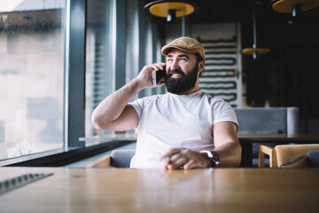 Cheerful caucasian mature male in casual wear enjoying rest in cafe interior talking on mobile phone, smiling bearded man 40s laughing during smartphone conversation satisfied with connection